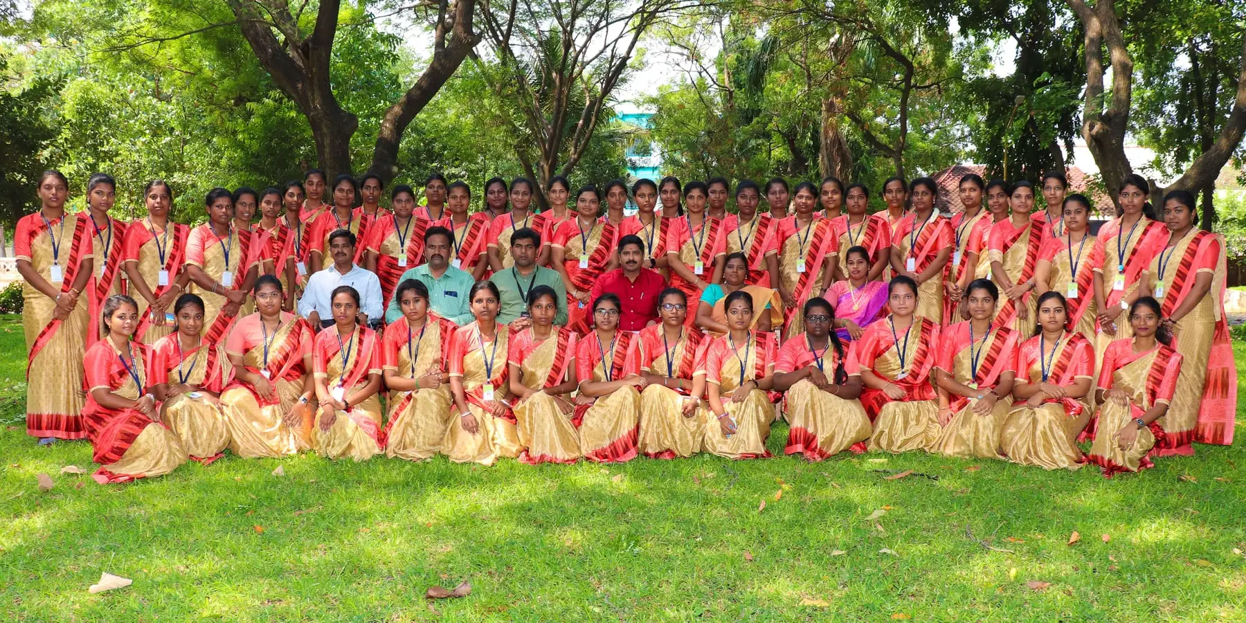 A batch of students in red and cream saris poses with faculty on a lawn under trees.