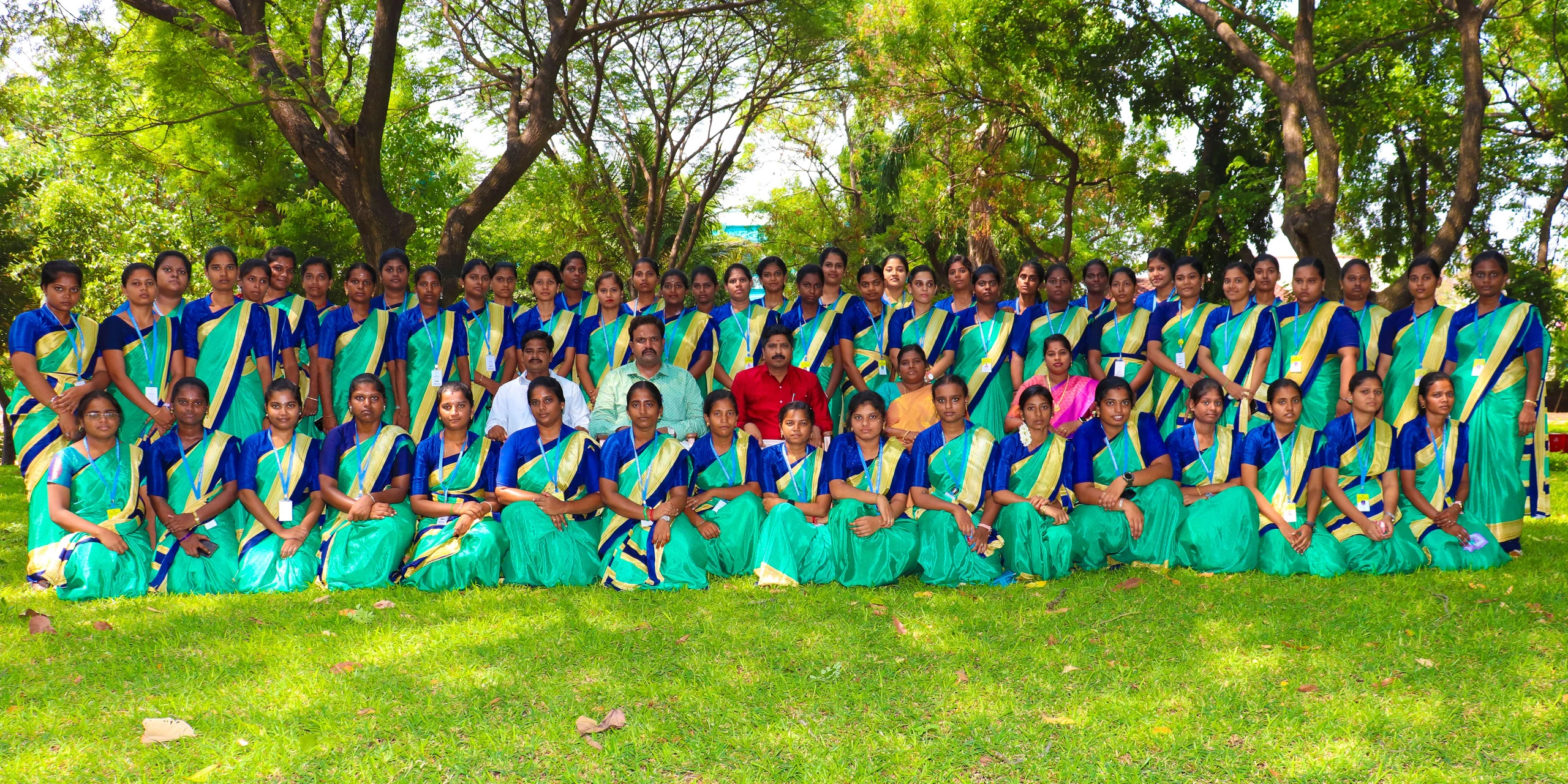 A batch of students in green and blue saris poses with faculty on a lawn under trees.