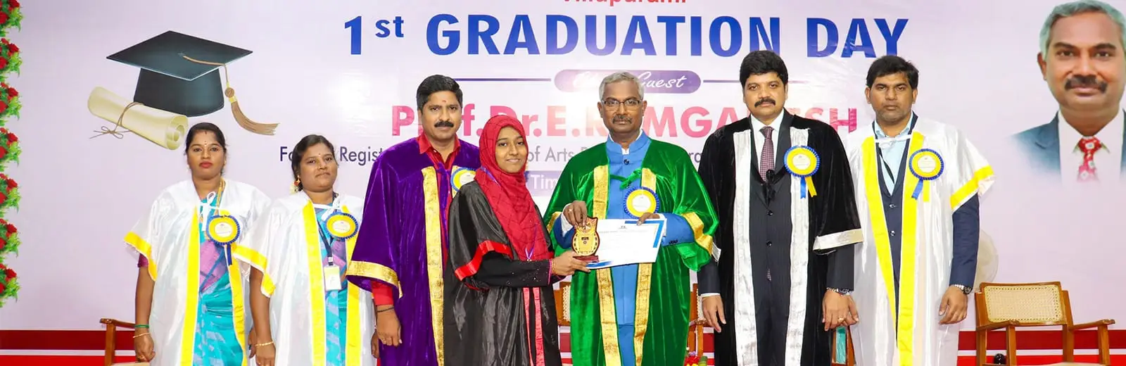A graduate in a gown receives her certificate on stage, under a "1st Graduation Day" banner.